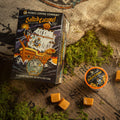 The Salted Caramel Bones Cups box is on top of a barrel with a canvas bag, caramel chunks, and a bones cup nearby. The art on the box shows a skeleton dressed as a pirate on a pirate ship with salted caramel on it.