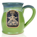 The front of the Bones Coffee Company Little Bounty Hunter Handthrown Mug. It has the Cantina Caramel Crunch art on the golden medallion. The mug is green colored with a black and white glaze.
