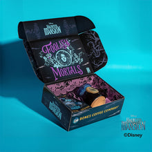 The collector's box inspired by Disney Haunted Mansion, it is laying on top of mossy rock. It is open and inside the box is a 12 ounce bag of flavored coffee named Brownie From Beyond, and beside the bag is a black colored mug with a purple glaze on it and a golden medallion in its center.