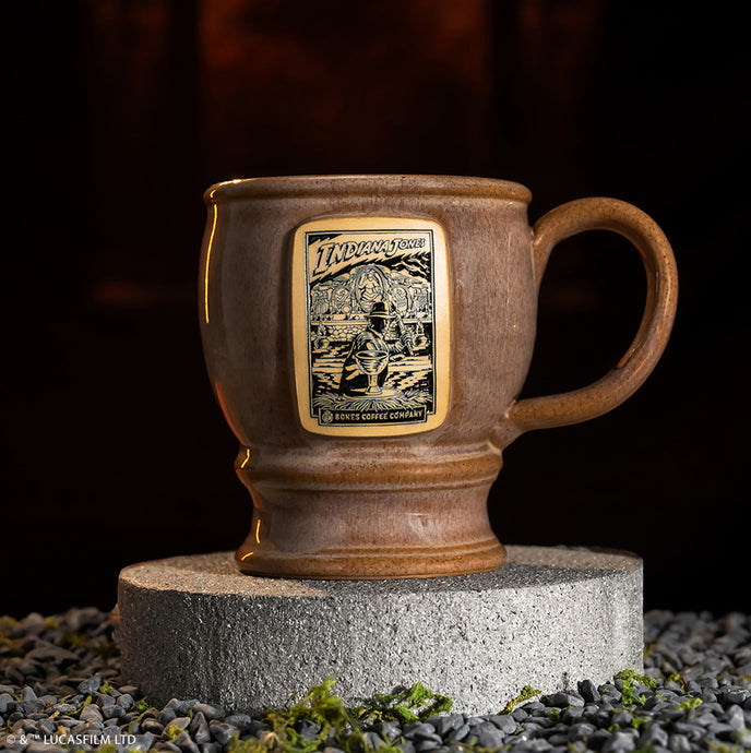 The front of the Bones Coffee Company Crusader’s Cup hand thrown mug with Indiana Jones on the golden medallion. It is inspired by Lucasfilm Indiana Jones. The mug is tan colored and on a stone pedestal.