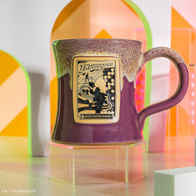 The front of the Bones Coffee Company Daring Delight hand thrown mug with Indiana Jones on the golden medallion. It is inspired by Lucasfilm Indiana Jones. The mug is purple colored and has a white glaze on top of it. It is set in a retro setting.