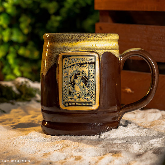 The front of the Bones Coffee Company Toffee Treasure hand thrown mug with Indiana Jones on the golden medallion. It is inspired by Lucasfilm Indiana Jones. The mug is brown colored and has a white glaze on top of it. It is sitting in front of a crate in sand.