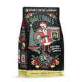 The front of a 12 ounce bag of Bones Coffee Company Jingle Bones coffee. Its flavor is coconut, caramel, and vanilla. It has a skeleton dressed as Santa and an elf in a living room with a Christmas tree on the art.