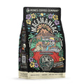 The front of a 12 ounce bag of Bones Coffee Company Macamaniac coffee. Its flavor is a coconut and macadamia nut, and it has a skeleton wearing a cap driving a red car sipping out of a coconut with an erupting volcano behind it on the art.