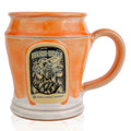 The front of the Bones Coffee Company Orange-Sickle hand thrown mug with the Orange-Sickle coffee art on the golden medallion. The mug is white colored with an orange glaze on top.