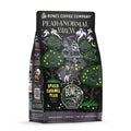 The front of a 12 ounce bag of Bones Coffee Company Pear-anormal Brew coffee. Its flavor is spiced caramel pear, and it has two skeletons walking through a haunted pear orchard towards a house full of ghosts on the art.