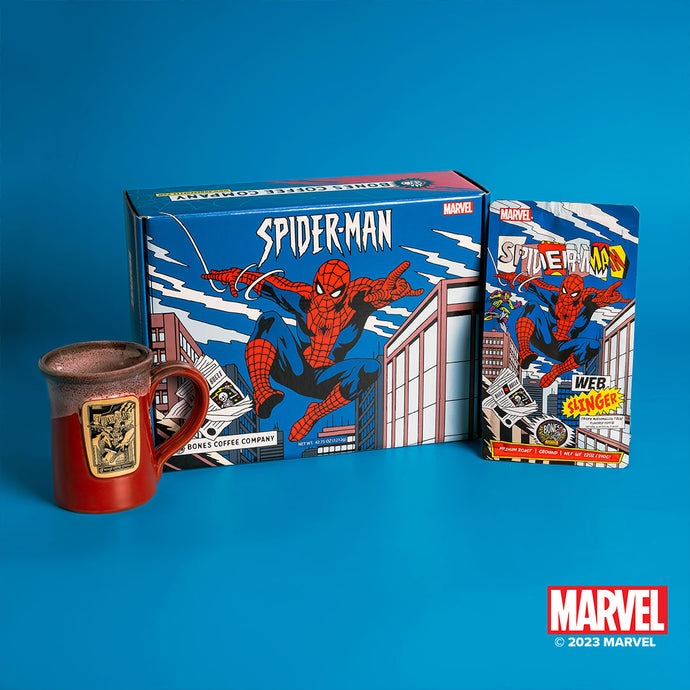 The collector's box inspired by Marvel Spider-Man. Around it is a 12 ounce bag of flavored coffee and a mug that is red colored with a white glaze on top of it with a golden medallion in the center.