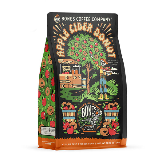 The front of a 12 ounce bag of Bones Coffee Company Apple Cider Donut coffee. Its flavor is apple cider donut, and it has a skeleton picking apples on the art. There is a sticker that says new flavor.