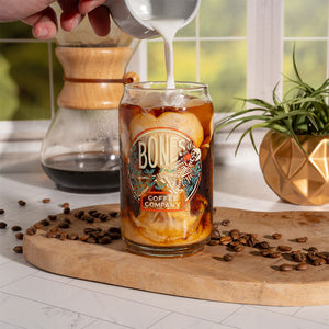 A cold brew coffee glass that has the Bones Coffee Company logo on it. The skeleton in the logo is sipping coffee and relaxing on greenery. Coffee and ice is inside the glass and someone is pouring cream into it.