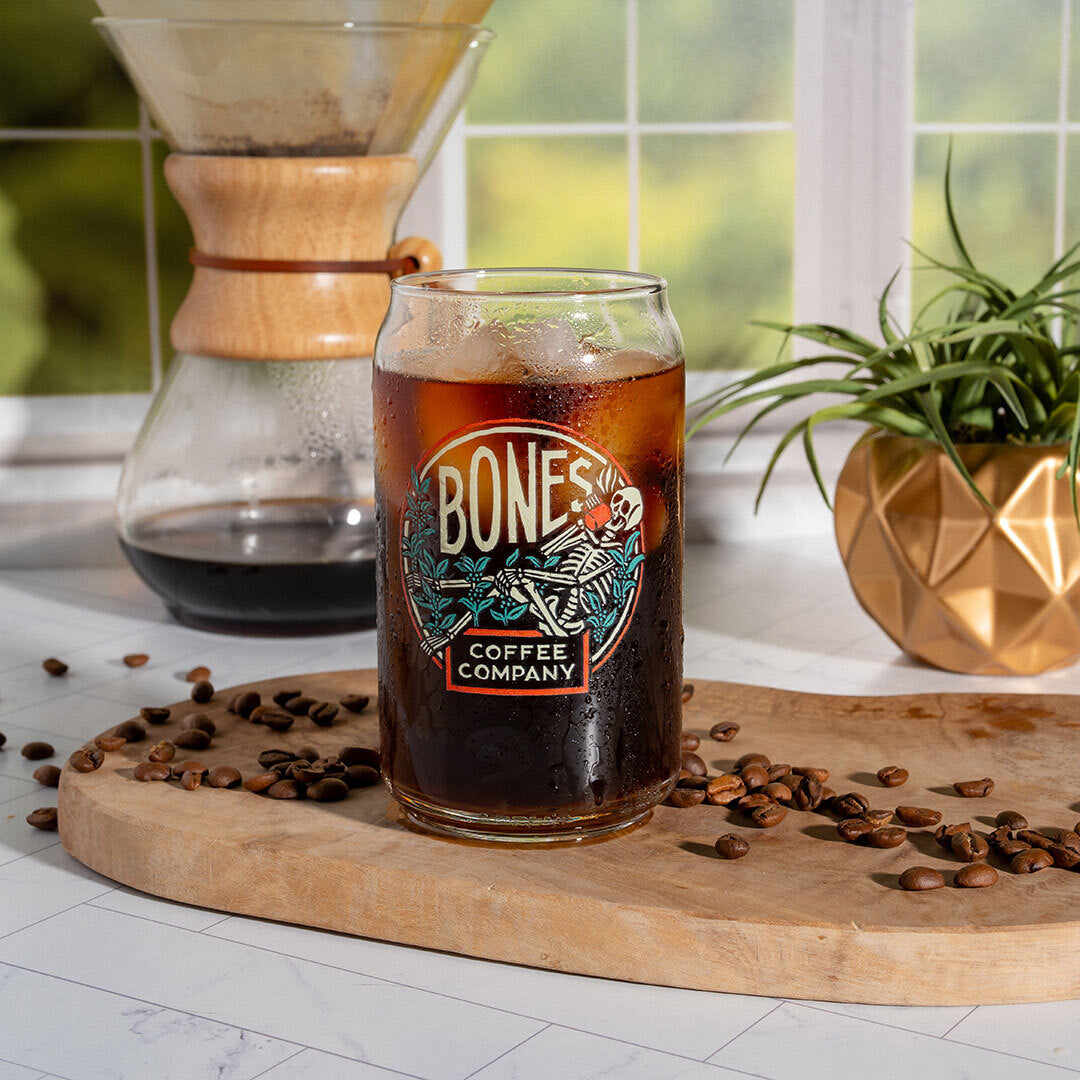 A cold brew coffee glass that has the Bones Coffee Company logo on it. The skeleton in the logo is sipping coffee and relaxing on greenery. Coffee and ice is inside the glass.