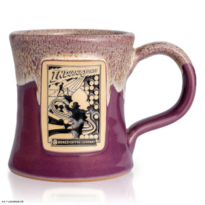 The front of the Bones Coffee Company Daring Delight hand thrown mug with Indiana Jones on the golden medallion. It is inspired by Lucasfilm Indiana Jones The mug is purple colored and has a white glaze on top of it.