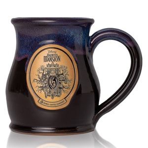 A mug inspired by Disney Haunted Mansion. It is colored black with a deep purple glaze on top of it with a golden medallion in the center holding art inspired by Disney Haunted Mansion.