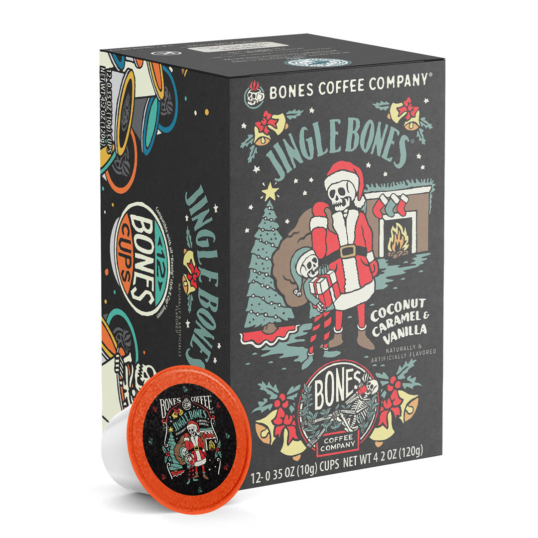 The front of the Bones Coffee Company Jingle Bones 12 Count Bones Cups box. Its flavor is coconut, caramel, and vanilla, and it has two skeletons dressed as Santa Clause and an elf standing inside a living room with a bag over the Santa skeleton’s shoulder on its art.