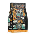 The front of a 12 ounce bag of Bones Coffee Company Maple Bacon coffee. Its flavor is bacon, and it has a skeleton dressed like a lumberjack sipping coffee in a forest with pigs on the art.
