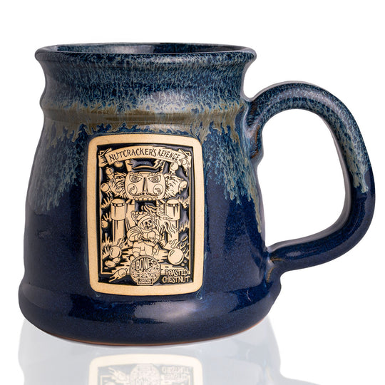 The front of the Bones Coffee Company Nutcracker’s Revenge hand thrown mug with the Nutcracker’s Revenge coffee art on the golden medallion. The mug is dark blue colored with a white glaze on top.