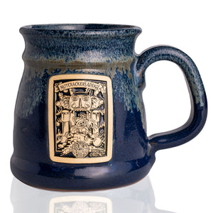 The front of the Bones Coffee Company Nutcracker’s Revenge hand thrown mug with the Nutcracker’s Revenge coffee art on the golden medallion. The mug is dark blue colored with a white glaze on top.