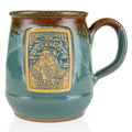 The front of the Bones Coffee Company Salty Siren Handthrown Mug. It has the Salty Siren art on the golden medallion. The mug is light teal colored with a cinnamon and white glaze.