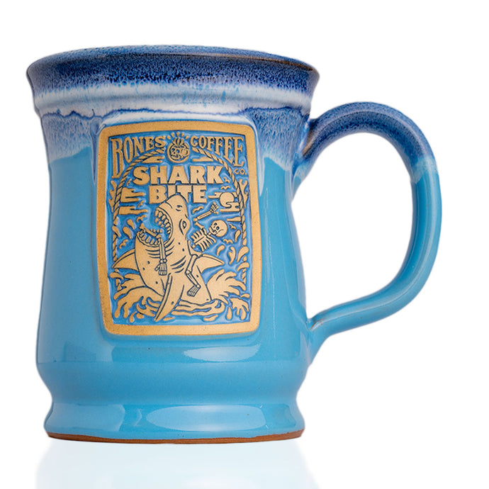 The front of the Bones Coffee Company Shark Bite hand thrown mug with the Shark Bite coffee art on the golden medallion. The mug is ocean blue colored with a white glaze on top.