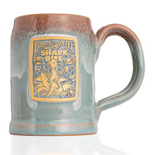 The front of the Bones Coffee Company Shark Bite hand thrown tankard with the Shark Bite coffee art on the golden medallion. The mug is blue colored with a cinnamon-white glaze on top.