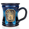 The front of the steamy stella mug. It is black colored with a blue glaze over it. The medallion shows a skeleton in a dress and heels inside a coffee mug with a leg up in the air.