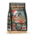 The front of a 12 ounce bag of Bones Coffee Company Toffee Almond Crunch coffee. Its flavor is toffee almond crunch, and it has a skeleton dressed up for christmas holding a basket of toffee almond crunch with a skeletal cat on the art.