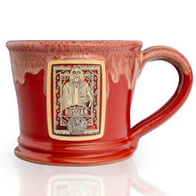 The front of the White Russian mug that is red colored with a sand-white glaze on top of it. There is a gold medallion on the center of the mug that has the art of White Russian coffee on it.