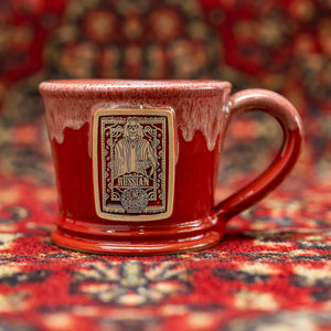 The front of the White Russian mug that is red colored with a sand-white glaze on top of it. There is a gold medallion on the center of the mug that has the art of What the Fluff coffee on it. The mug is on a rug.