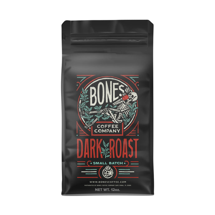 The front of a 12 ounce coffee bag of Bones Coffee Company Dark Roast coffee. It has the Bones Coffee Company logo that has a skeleton sipping coffee, lounging on greenery around on it on the art.