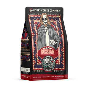 The front of a 12 ounce bag of Bones Coffee Company White Russian coffee. Its flavor is cream and coffee liqueur, and it has a skeleton wearing sunglasses with a goatee in a comfy coat with a white tee shirt holding a cup of coffee on the art.