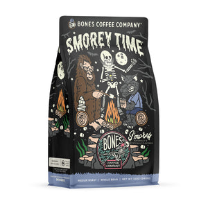 The front of a 12 ounce bag of Bones Coffee Company S’morey Time coffee. Its flavor is s’mores, and it has a skeleton holding a flashlight around a campfire with bigfoot and a werewolf on the art.
