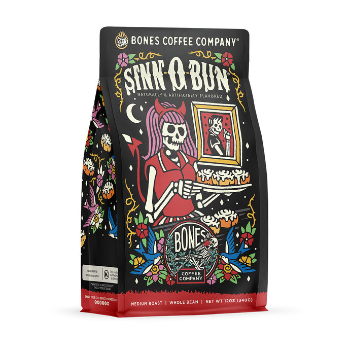 The front of a 12 ounce bag of Bones Coffee Company Sinn-O-Bun coffee. Its flavor is cinnamon bun, and it has a skeleton with pink hair wearing a horned headband holding a tray of cinnamon buns on the art.