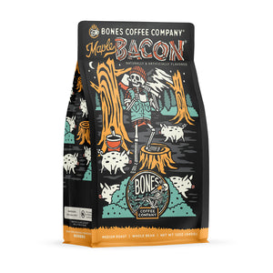 The front of a 12 ounce bag of Bones Coffee Company Maple Bacon coffee. Its flavor is bacon, and it has a skeleton dressed like a lumberjack sipping coffee in a forest with pigs on the art.