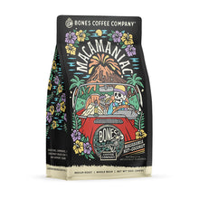 The front of a 12 ounce bag of Bones Coffee Company Macamaniac coffee. Its flavor is a coconut and macadamia nut, and it has a skeleton wearing a cap driving a red car sipping out of a coconut with an erupting volcano behind it on the art.