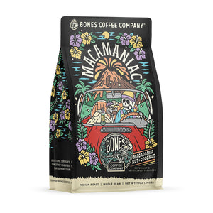 The front of a 12 ounce bag of Bones Coffee Company Macamaniac coffee. Its flavor is a coconut and macadamia nut, and it has a skeleton wearing a cap driving a red car sipping out of a coconut with an erupting volcano behind it on the art.