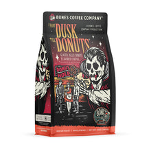 The front of a 12 ounce bag of Bones Coffee Company From Dusk til Dawn coffee. Its flavor is jelly donut, and it has a skeleton dressed like a vampire holding a jelly donut while people leave a donut shop on the art.