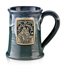 The front of the Salty Siren mug that is seafoam colored with an ocean blue glaze on top of it. There is a gold medallion on the center of the mug that has the art from the Salty Siren coffee on it.