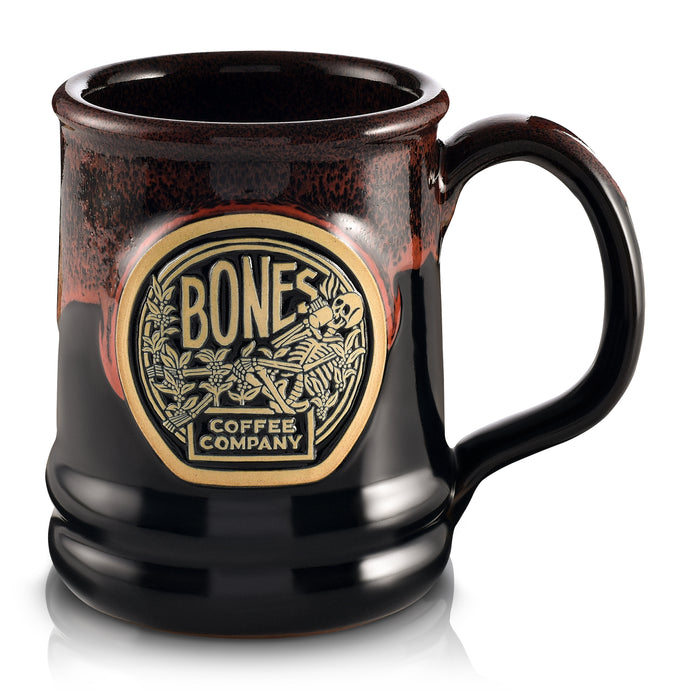The front of the Bones Coffee Company Classic Logo mug that is black colored with an orange glaze on top of it. There is a gold medallion on the center of the mug that has the classic logo for Bones Coffee Company of a skeleton sipping coffee on top of greenery on it.