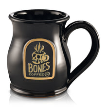 The front of the Bones Coffee Company Skull Logo mug that is black colored. There is a gold medallion on the center of the mug that has the skull logo for Bones Coffee Company of a skull that looks like a mug with steam on top of it on it.