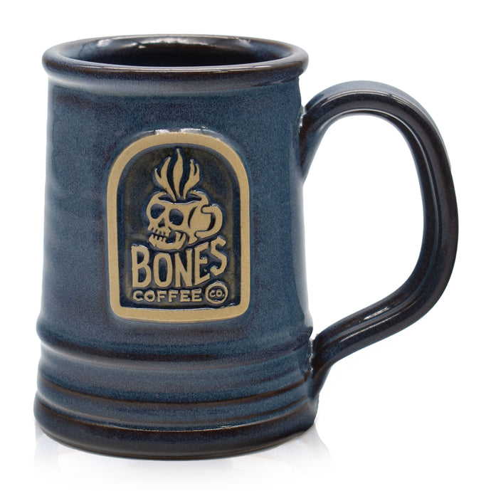 The front of a denim colored mug with the Bones Coffee Company logo on it in a golden medallion. Above the words Bones Coffee Co is a skull that looks like a mug and has steam coming out of the top of it.
