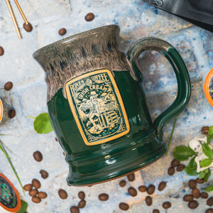 The front of the Bones Coffee Company Irish Cream hand thrown tankard with the Irish Cream coffee art on the golden medallion. The mug is green colored and has a coffee glaze on top of it. It is on a stone surface with coffee beans.