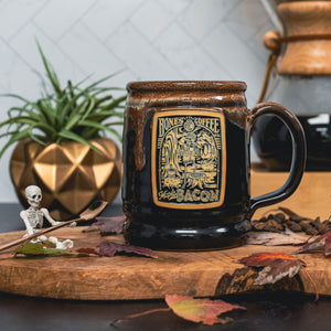 The front of the Bones Coffee Company Maple Bacon hand thrown mug with the Maple Bacon coffee art on the golden medallion. The mug is black colored with a bacon glaze on top. It is on a table with a small skeleton.