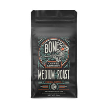 The front of a 12 ounce coffee bag of Bones Coffee Company Medium Roast coffee. It has the Bones Coffee Company logo that has a skeleton sipping coffee, lounging on greenery around on it on the art.