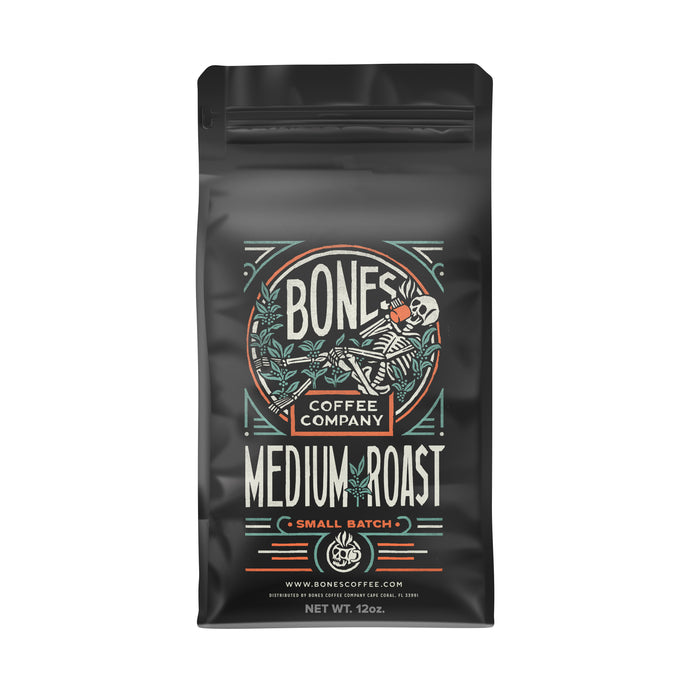 The front of a 12 ounce coffee bag of Bones Coffee Company Medium Roast coffee. It has the Bones Coffee Company logo that has a skeleton sipping coffee, lounging on greenery around on it on the art.