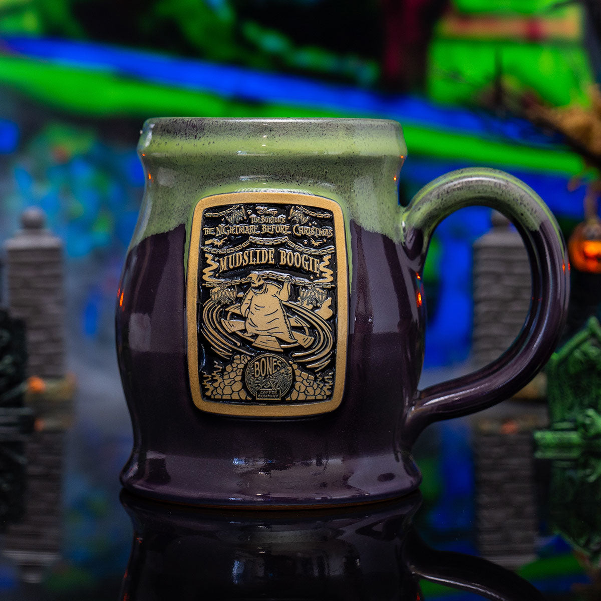 The front of the Bones Coffee Company Mudslide Boogie hand thrown mug with Oogie Boogie on the golden medallion. It is inspired by Disney Tim Burton’s The Nightmare Before Christmas. The mug is plum colored with a kiwi green glaze on top. It is inside a toy graveyard.