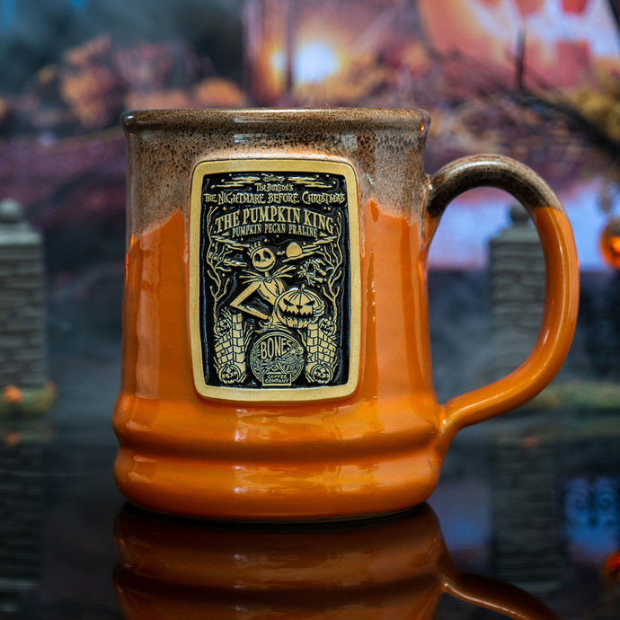 The front of the Bones Coffee Company The Pumpkin King hand thrown mug with Jack Skellington on the golden medallion. It is inspired by Disney Tim Burton’s The Nightmare Before Christmas. The mug is orange colored with a black and white glaze on top. It is inside a toy graveyard.
