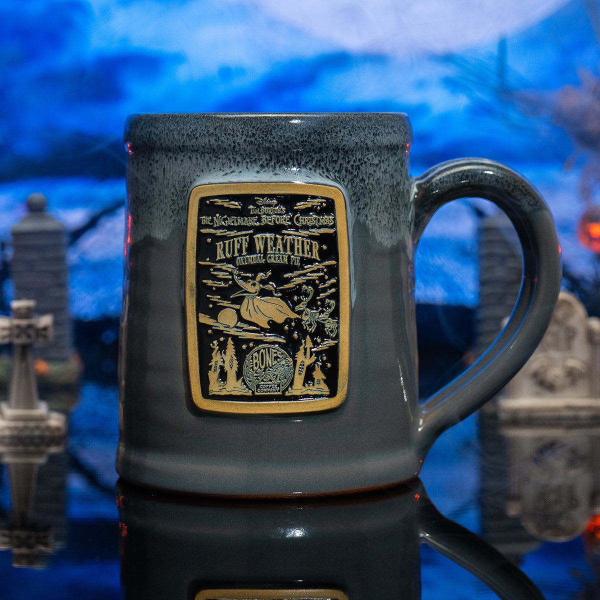 The front of the Bones Coffee Company Ruff Weather hand thrown mug with Zero the dog on the golden medallion. It is inspired by Disney Tim Burton’s The Nightmare Before Christmas. The mug is grey colored with a black and white glaze on top. It is inside a toy graveyard.