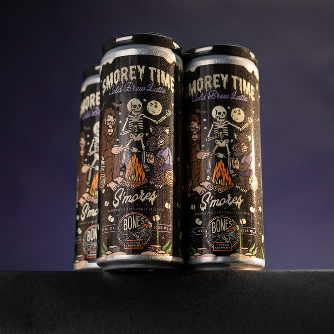 A four pack of S'morey Time cold brew lattes. The art on the can shows a skeleton holding a flashlight around a campfire with bigfoot and a werewolf.