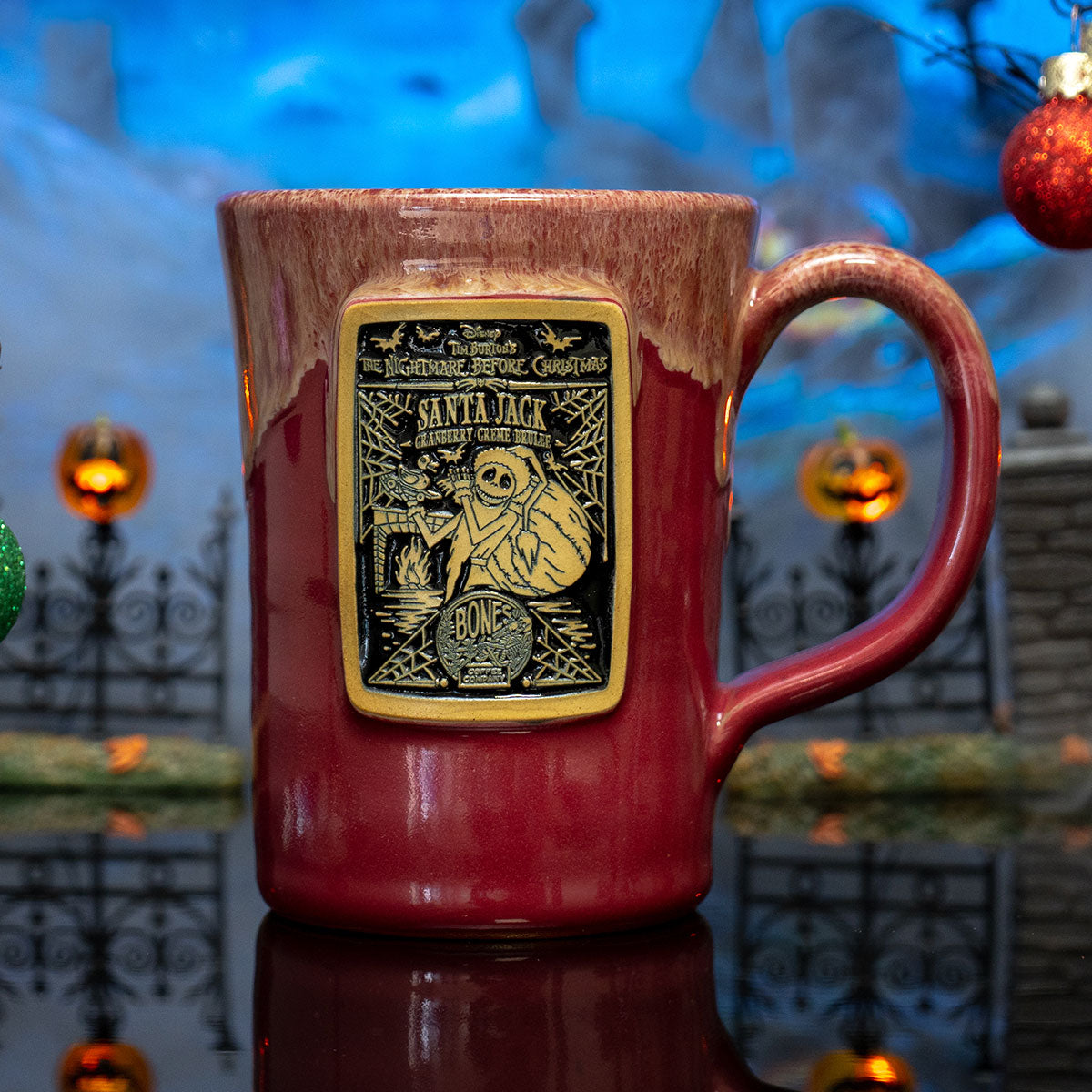 The front of the Bones Coffee Company Santa Jack hand thrown mug with Jack Skellington on the golden medallion. It is inspired by Disney Tim Burton’s The Nightmare Before Christmas. The mug is cranberry colored with a white glaze on top. It is inside a toy graveyard.