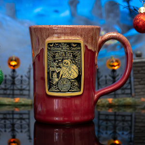The front of the Bones Coffee Company Santa Jack hand thrown mug with Jack Skellington on the golden medallion. It is inspired by Disney Tim Burton’s The Nightmare Before Christmas. The mug is cranberry colored with a white glaze on top. It is inside a toy graveyard.