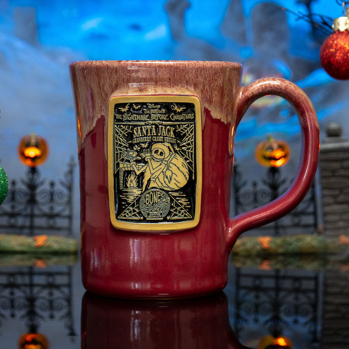 The front of the Bones Coffee Company Santa Jack hand thrown mug with Jack Skellington on the golden medallion. It is inspired by Disney Tim Burton’s The Nightmare Before Christmas. The mug is cranberry colored with a white glaze on top. It is inside a toy graveyard.